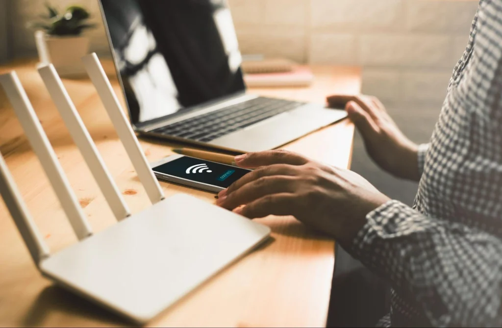 Personne vérifiant la connexion Wi-Fi sur un smartphone, assise près d'un routeur et d'un ordinateur portable.