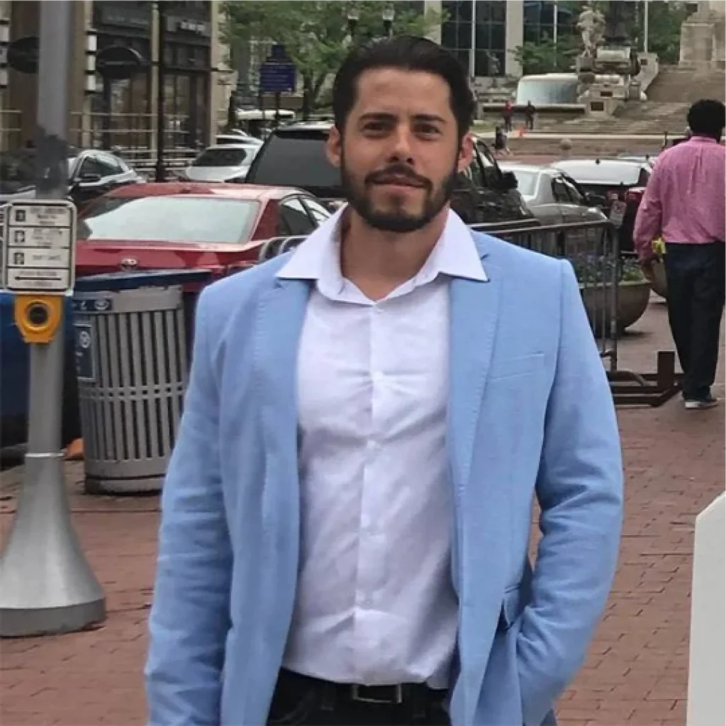 Jeff tavares, vêtu d'un blazer bleu clair et d'une chemise blanche, se tient à l'extérieur, avec des voitures et des bâtiments en arrière-plan.