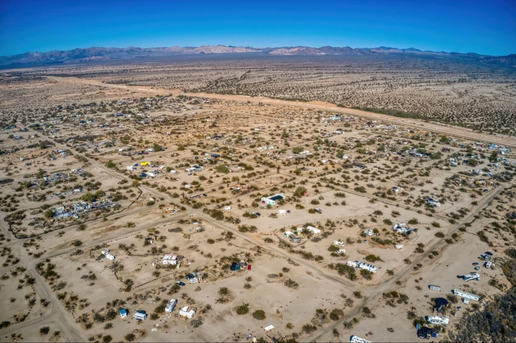 Vue aérienne de l'agglomération désertique de Slab City, avec des véhicules de loisirs et des habitations de fortune éparpillés, sous un ciel bleu.