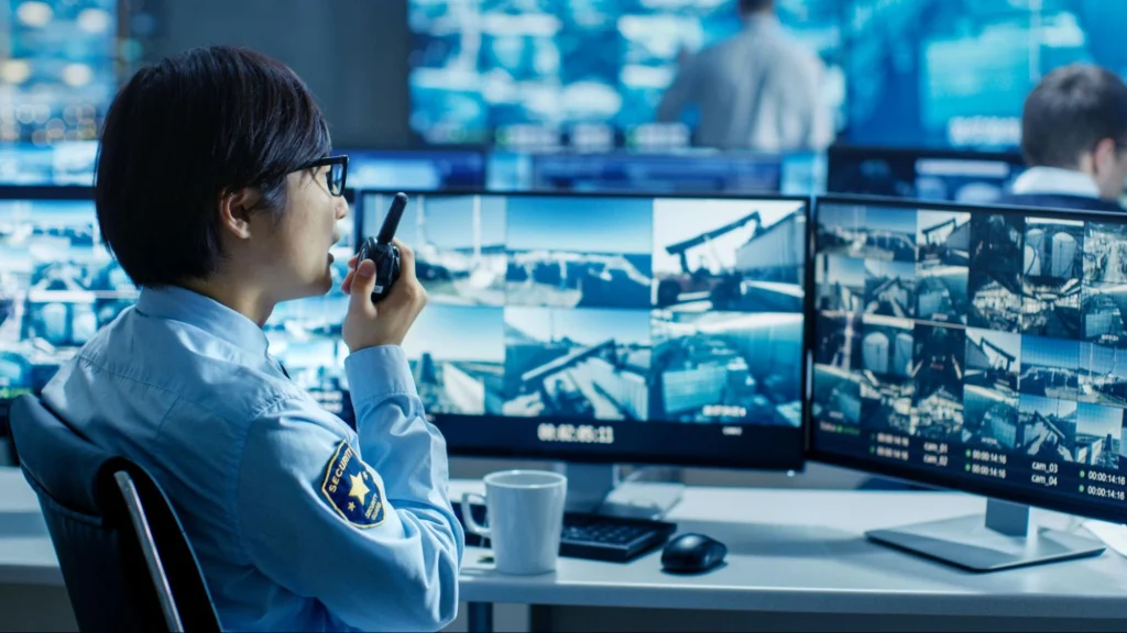 Agent de sécurité contrôlant plusieurs caméras de surveillance sur des écrans tout en parlant dans un talkie-walkie.